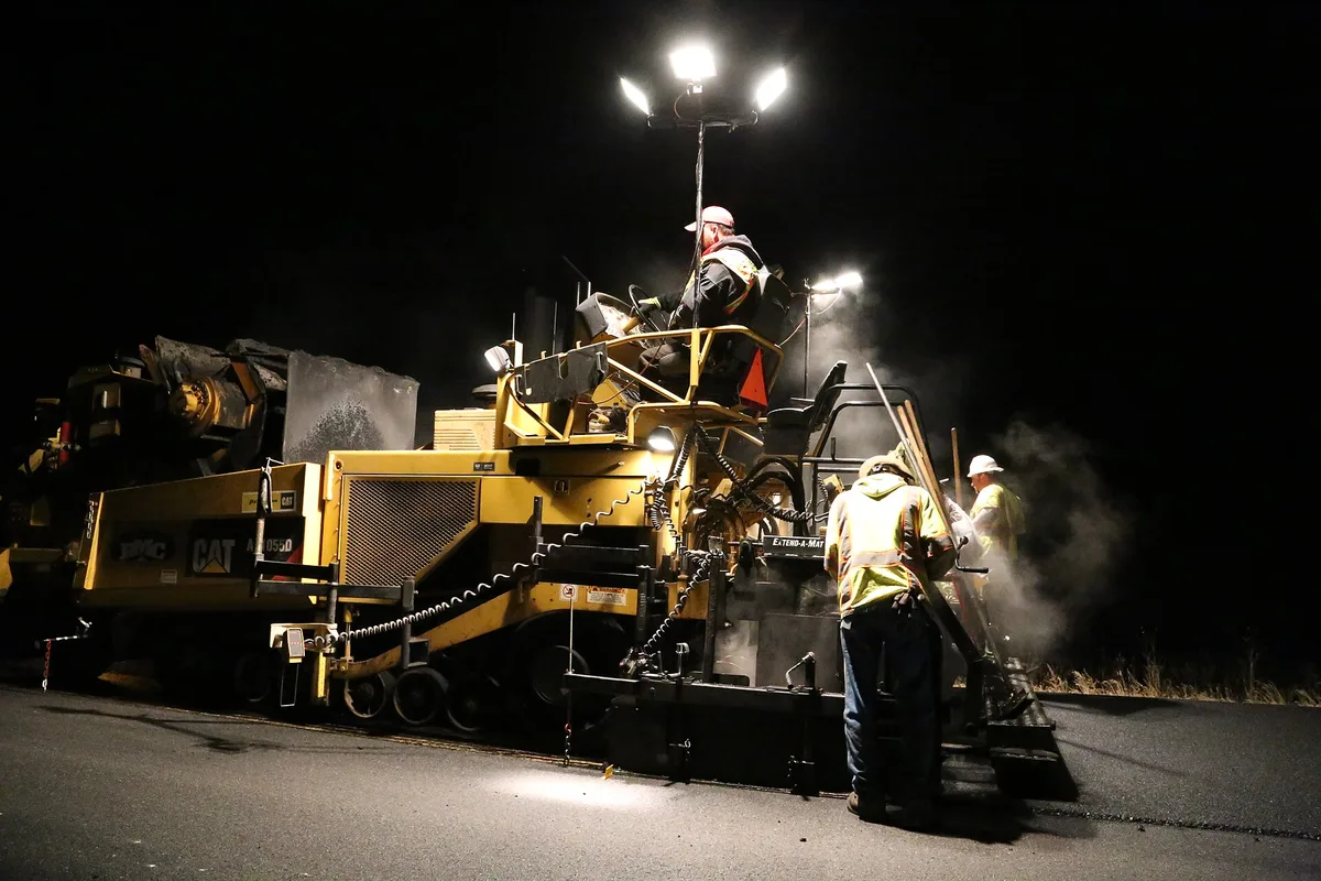 Road crew in safety vests operating a paving machine under work lights at night